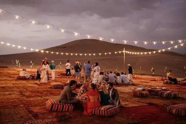 Grupo sentado sobre alfombras y cojines bajo guirnaldas de luces, desierto de Agafay